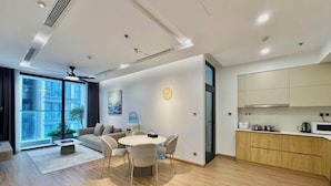 A modern living room with a round table and chairs, a sofa, a large window, and a kitchen area with cabinets and appliances. - Vinhomes Metropolis Apartment - Comfort Stay Ha Noi (Hanoi)