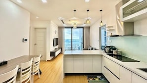 A modern kitchen with white cabinets, a dining area with a wooden table and chairs, and a large window with curtains. - Vinhomes Metropolis Apartment - Comfort Stay Ha Noi (Hanoi)