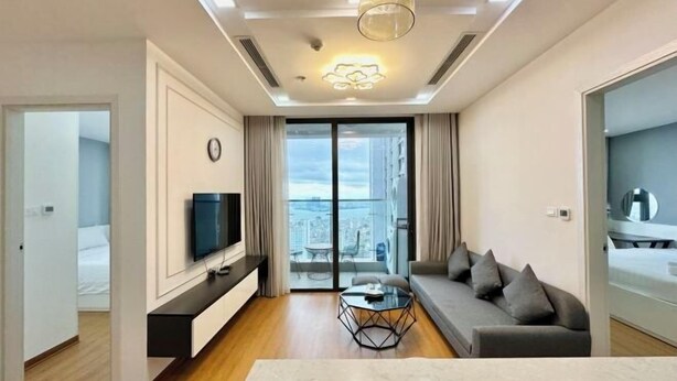 A modern living room with a flat-screen TV, a sofa, a coffee table, and a balcony with a city view. - Vinhomes Metropolis Apartment - Comfort Stay Ha Noi (Hanoi)