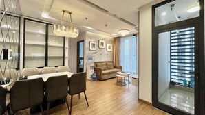 A modern living room with a dining area, a sofa, a chandelier, and a large window with blinds. - Vinhomes Metropolis Apartment - Comfort Stay Ha Noi (Hanoi)