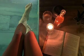 A person relaxing in a bathtub with a book and candle on a table.