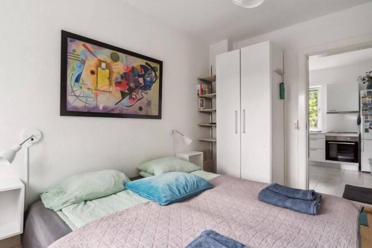 A modern bedroom with a bed, pillows, a painting, a wardrobe, and a desk with a lamp.