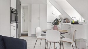 A modern kitchen with white cabinetry, a dining area with a round table and chairs, and a visible oven. - Ama Hyg Atelier (Copenhagen)