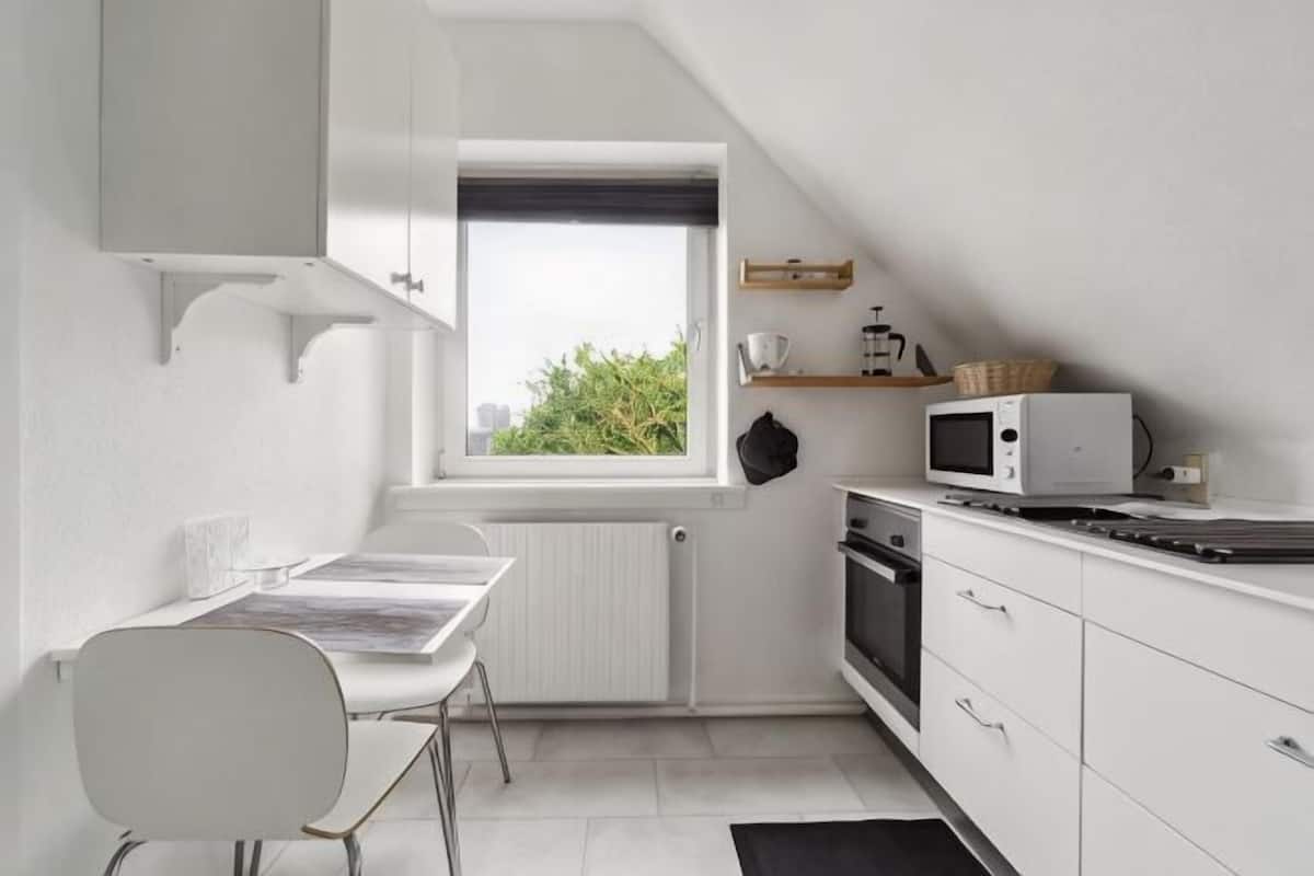 Eine kleine Küche mit Esstisch, weißen Schränken, einer Mikrowelle und einem Fenster mit Blick auf Grünflächen.