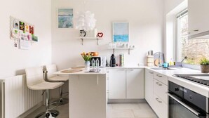 A modern kitchen with white cabinetry, a central island, bar stools, and a view of the outdoors through a window. - Ama Hyg Atelier (Copenhagen)