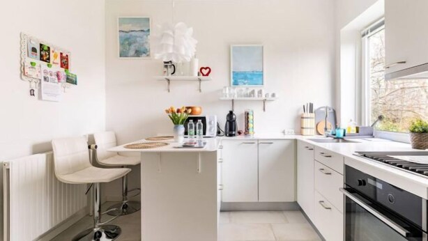 A modern kitchen with white cabinetry, a central island, bar stools, and a view of the outdoors through a window. - Ama Hyg Atelier (Copenhagen)