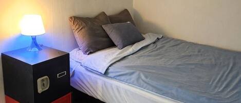 Uma cama de solteiro com colchão azul, uma mesa de cabeceira com abajur e um móvel de armazenamento vermelho.