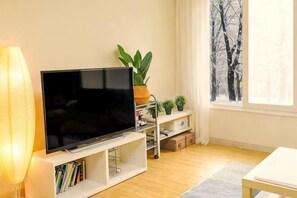 A modern living room with a flat-screen TV, a bookshelf, a plant, and a window with a view of trees. - Mark's House(8 minutes from Hongik Station/3Room/4Bed) (Seoul)