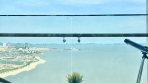 A view from a window with a telescope, overlooking a coastal area with buildings and a beach. - Alacarte Ha Long - Service Apartment (Ha Long)