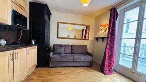 A compact living space with a kitchenette, a sofa, a large mirror, and a window with a curtain. - Vintage studio 150m to Notre Dame (Paris)