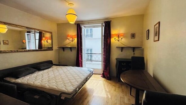 A hotel room with a bed, a desk, a chair, a mirror, two wall-mounted lamps, a window with curtains, and a view of the outside. - Vintage studio 150m to Notre Dame (Paris)