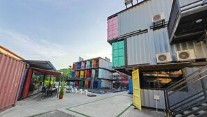 Μια αυλή με στοιβάζοντα μεταλλικά κοντέινερ μεταφοράς, μερικά από τα οποία διαθέτουν μονάδες κλιματισμού, και μια μικρή εξωτερική περιοχή με τραπέζια και καρέκλες για να καθίσουν οι άνθρωποι.