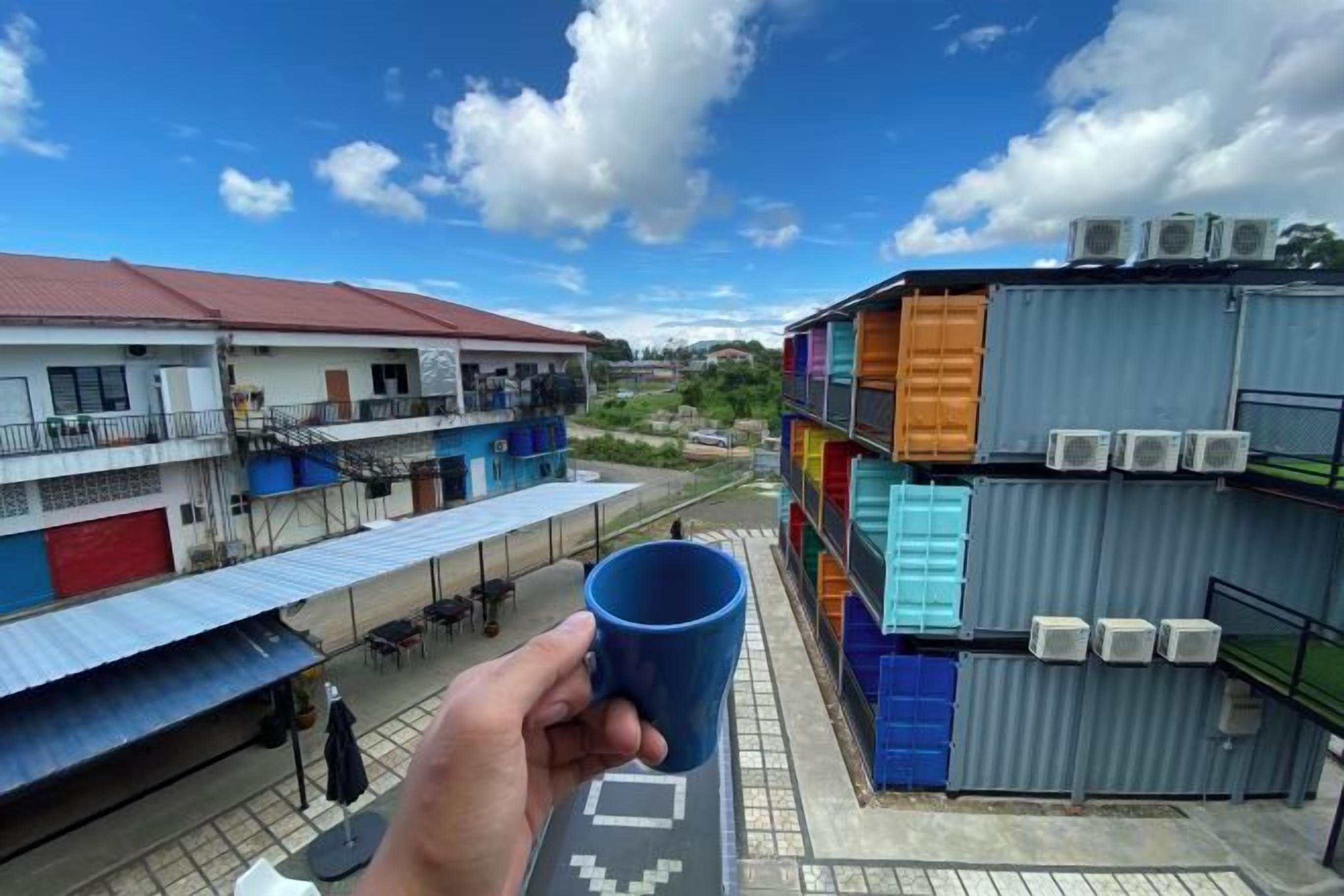 A hand holding a cup, overlooking a building with multiple floors and balconies, and a clear sky with clouds.