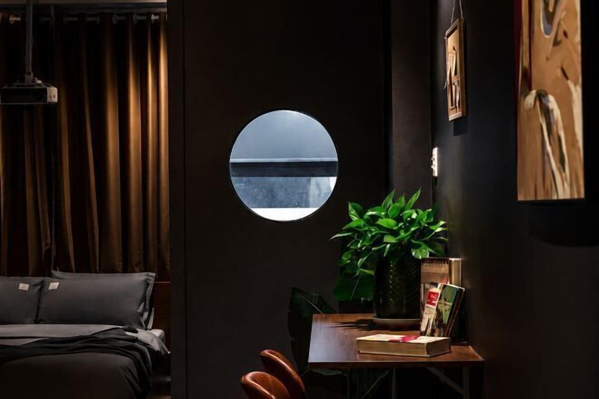 A room with a bed, a round window, a plant, a table with books, and a painting on the wall.