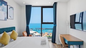 Habitación de hotel con cama, escritorio, silla y un amplio ventanal con vista al mar.