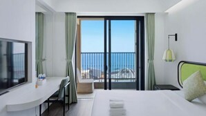 Una habitación de hotel con cama, escritorio, televisión, balcón con vista al mar y una lámpara.