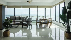 A spacious room with a large window offering a view of the sea, a ceiling fan, a sofa, a dining table with chairs, and two potted plants.