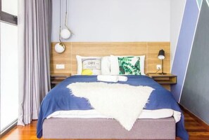 A neatly made bed with a blue and white bedspread, a white throw blanket, and two pillows, one with a green leaf pattern. There is a nightstand with a lamp and a power outlet on the wall beside the bed. - 270° Seaview, Rumahku Gurney (George Town)