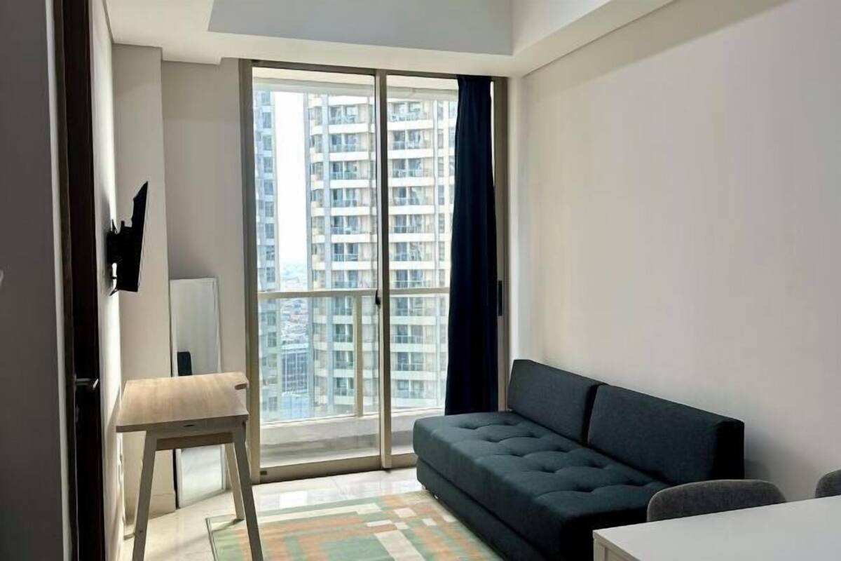 A modern living room with a sofa, a small table, and a view of high-rise buildings through a large window.