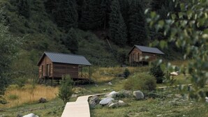 Dos cabañas de madera con techos metálicos, un sendero de madera y una ladera cubierta de bosque.