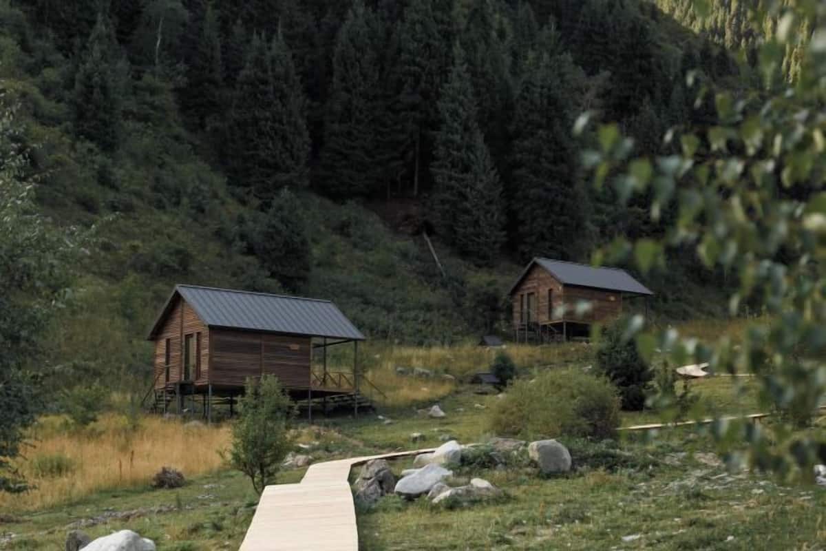 Deux cabanes en bois avec des toits en métal, un sentier en bois et une colline boisée.