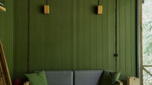 Una habitación con un muro verde paneado, un sofá gris con cojines y un ventanal con vistas a la vegetación.