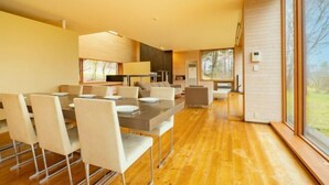 A modern dining area with a long table and chairs, a kitchen in the background, and large windows offering a view of the outdoors. - Toyako Villa Susukian (Usu District)