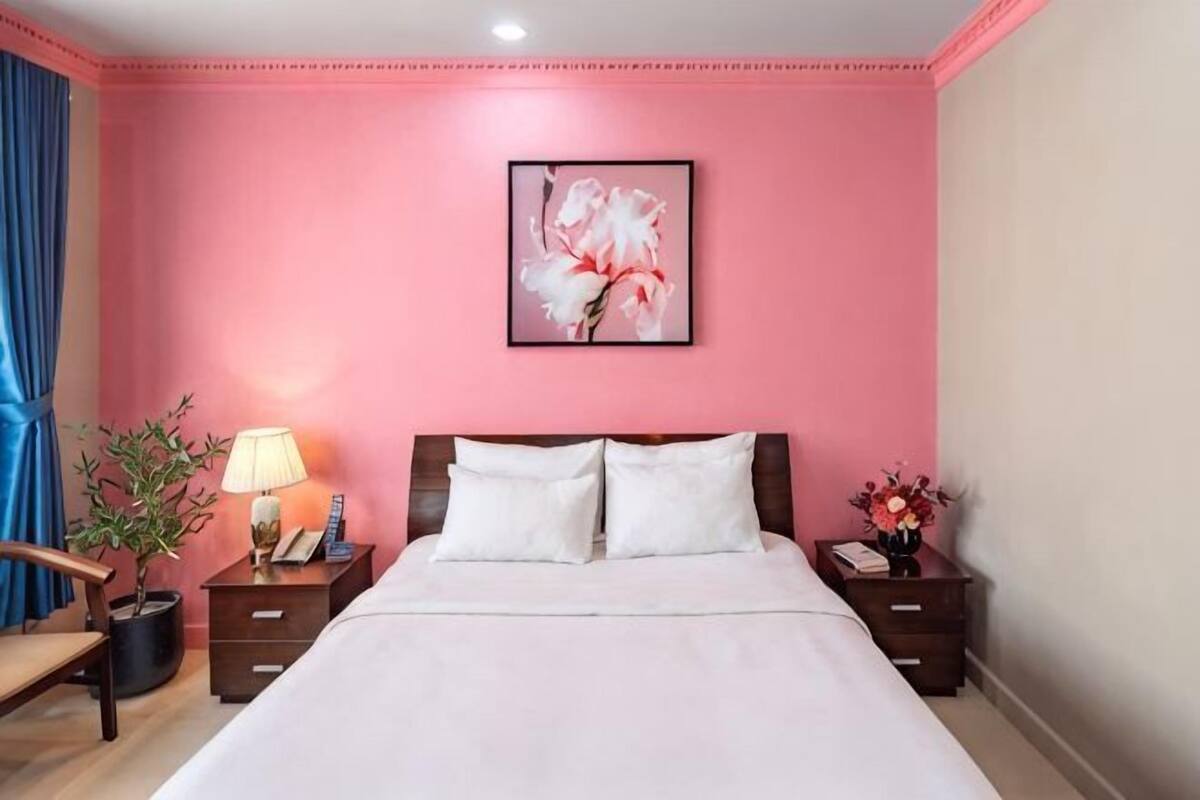 A hotel room with a pink wall, a bed with white bedding, a nightstand with a lamp, and a framed picture above the bed.