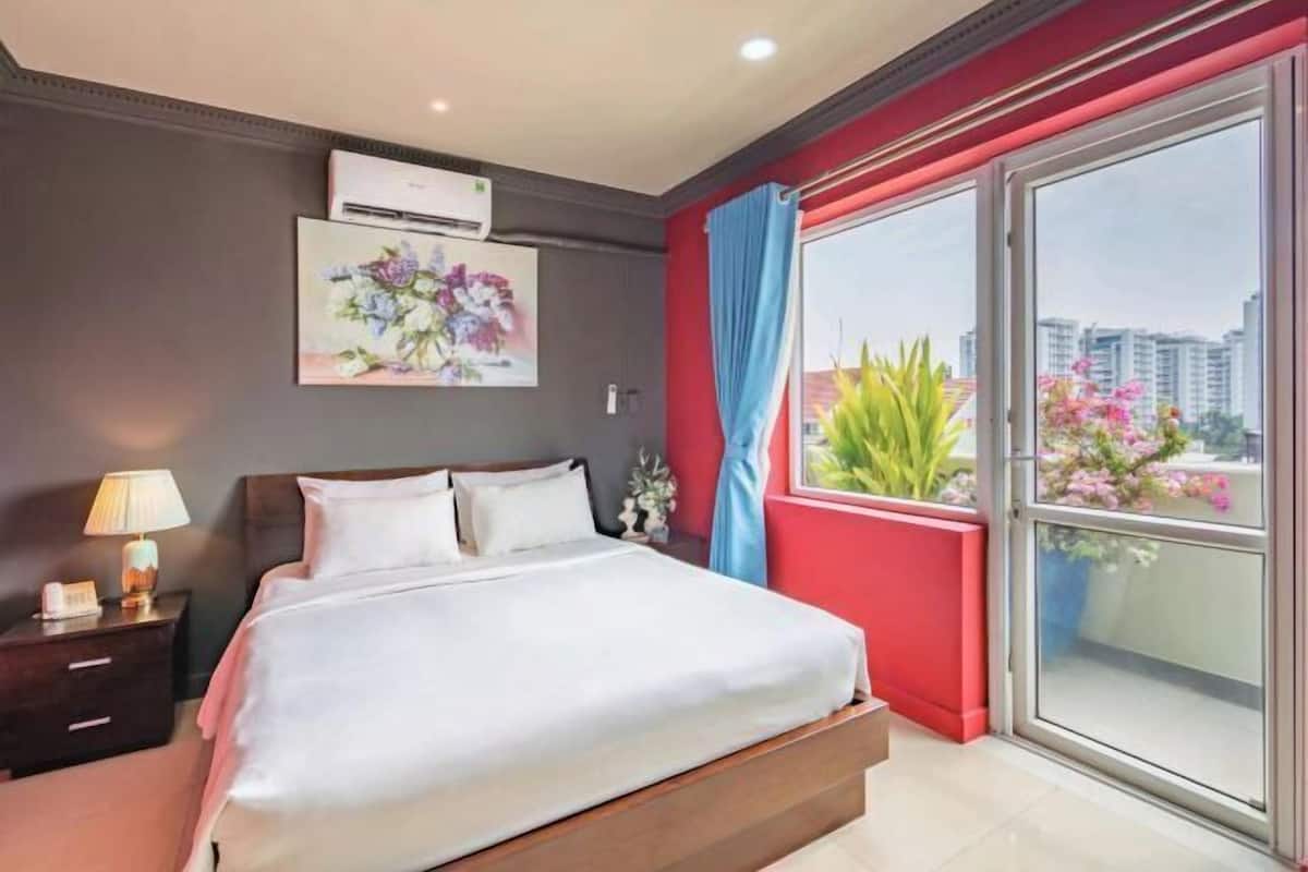 A hotel room with a bed, a nightstand, an air conditioning unit, a window with a view of buildings and plants, and a red curtain.