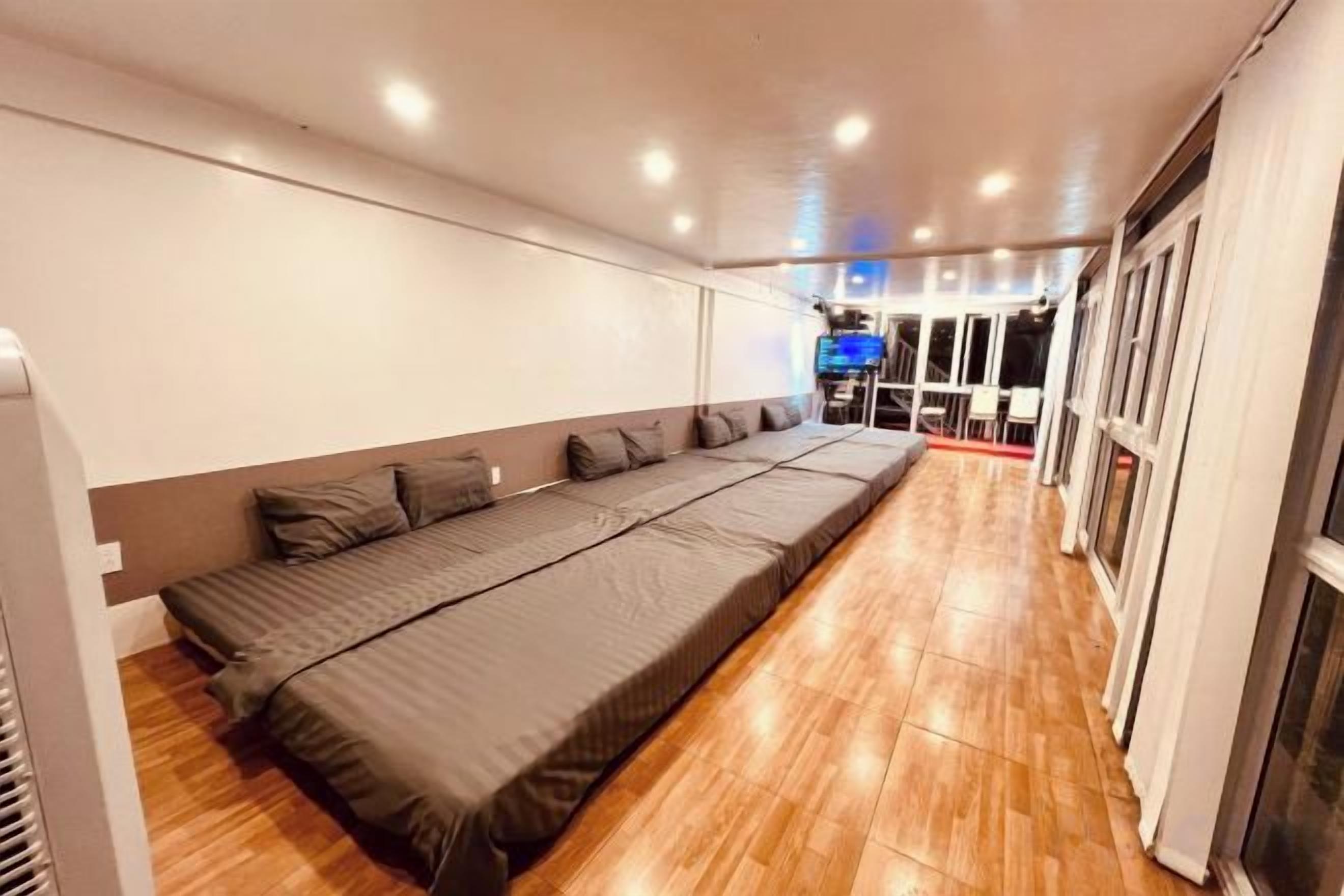 A room with a long bed, wooden flooring, and a television on the far wall.