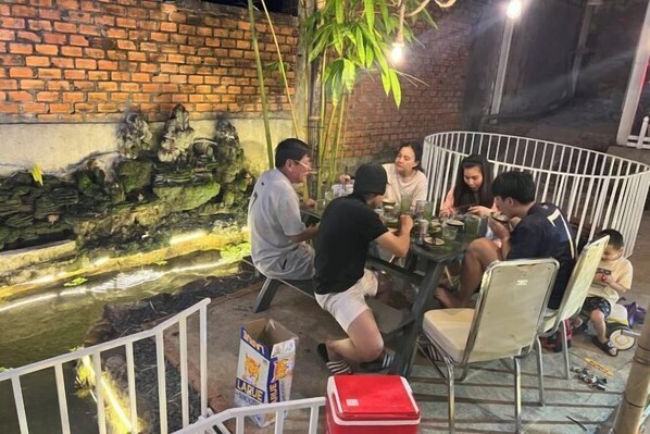 A group of people dining at an outdoor table with a brick wall and a decorative pond in the background. - Nice Hotel Buon Ma Thuot (Tu An)