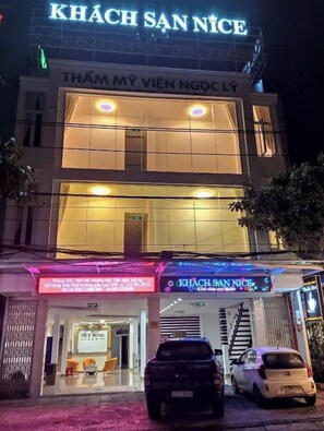 Un hôtel appelé « KHÁCH SAN NICE », doté d’une façade moderne, d’une entrée bien visible et d’un parking où sont garées des voitures.