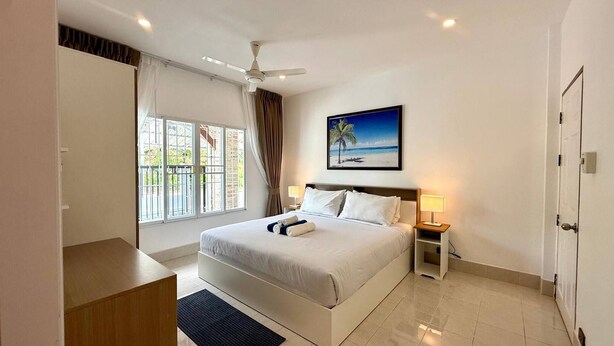 A hotel room with a bed, a desk, a chair, a ceiling fan, a window with curtains, a nightstand with a lamp, and a painting of a beach scene.