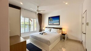 A hotel room with a bed, a desk, a chair, a ceiling fan, a window with curtains, a nightstand with a lamp, and a painting of a beach scene.