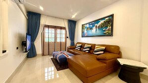 A living room with a brown leather sofa, a small ottoman, a television, and a painting of a tropical landscape.