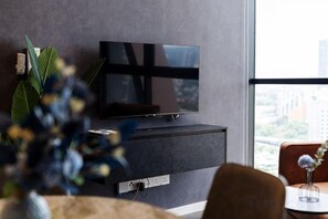 A modern living room with a flat-screen TV on a dark cabinet, a large window with a view of buildings, and a plant in the foreground. - SCARLETZ BY FEEL HOME (Kuala Lumpur)