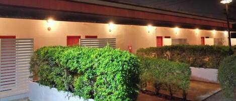 Un edificio con varias entradas, fachada iluminada y zona ajardinada con arbustos.