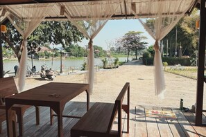 A covered outdoor seating area with wooden tables and benches, overlooking a lake and a garden. - Ingnaam Thajeen (Si Prachan)