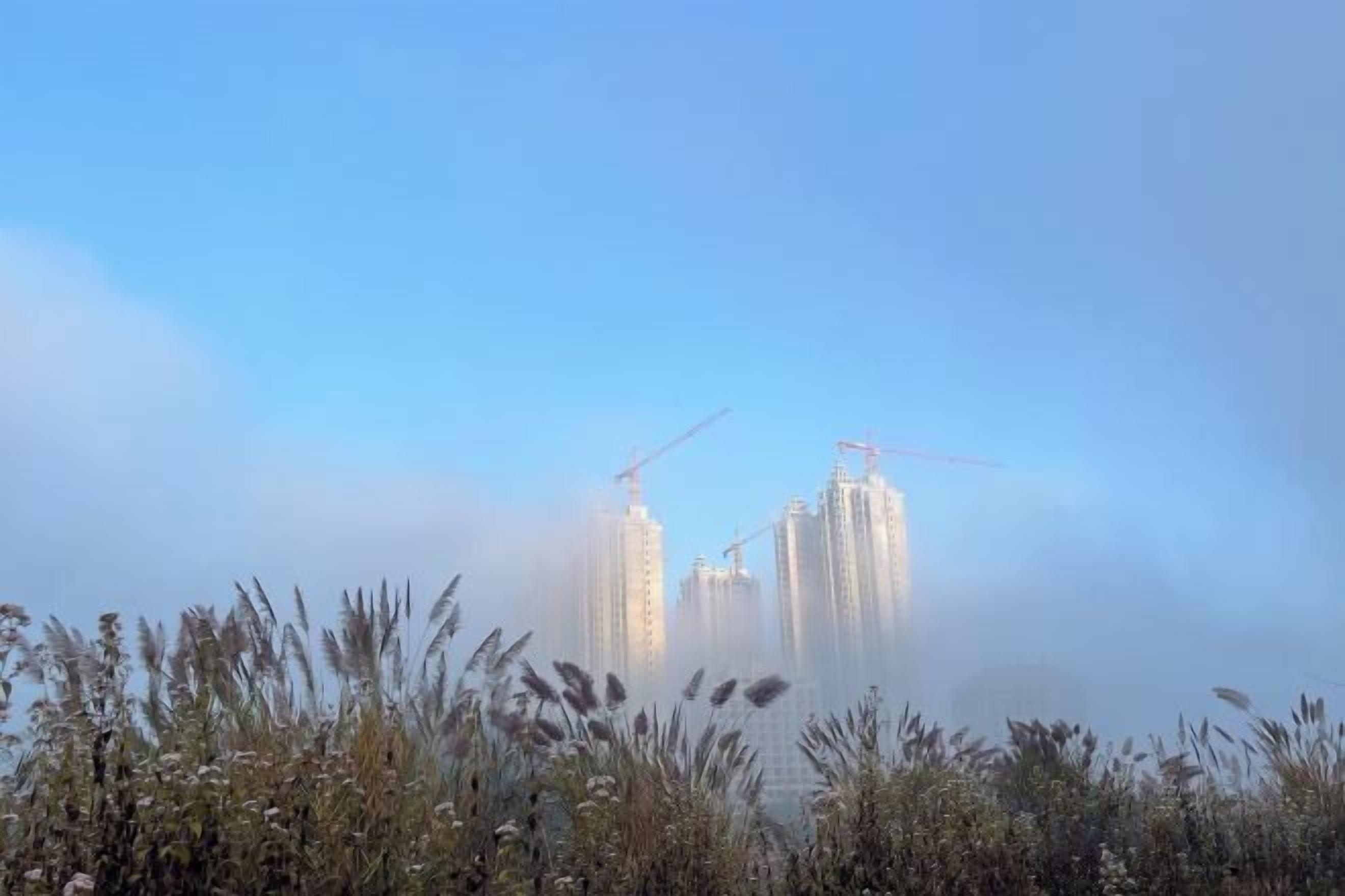 A high-rise building with construction cranes, surrounded by fog.