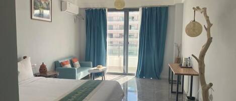 A hotel room with a bed, a sofa, a small table, a chair, a window with curtains, and a balcony.