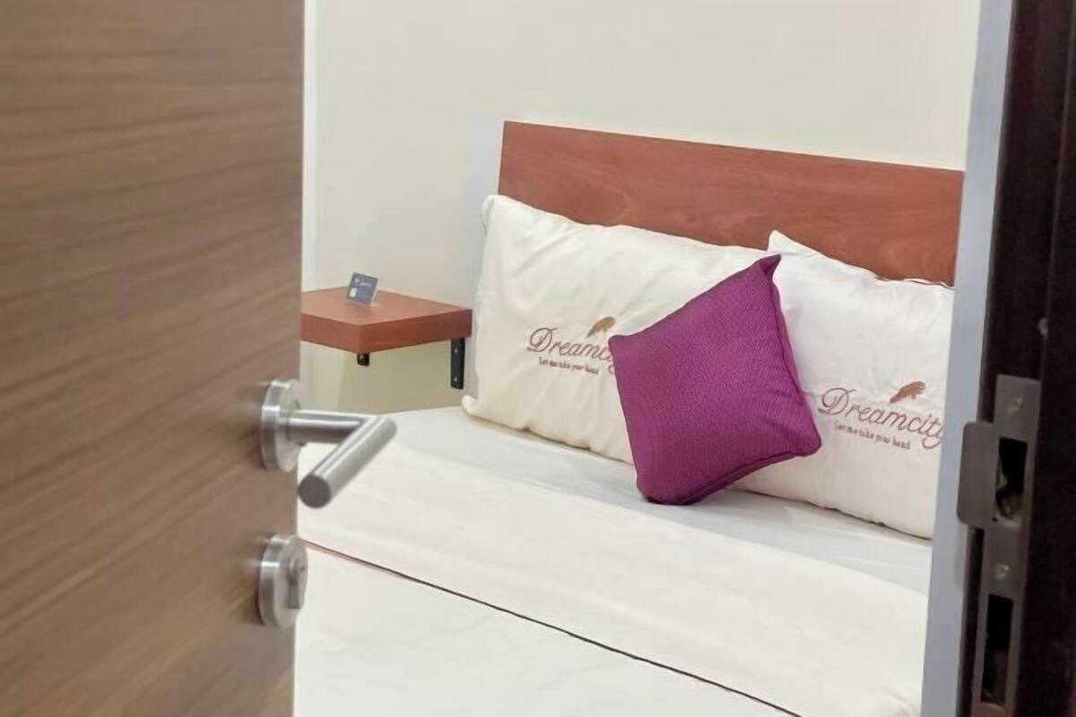Ein kleines Bett mit Holz-Kopfteil, zwei Kissen und ein Nachttischchen mit einer Lampe.