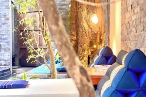A cozy seating area with cushioned chairs and a table, surrounded by stone walls and a tree.