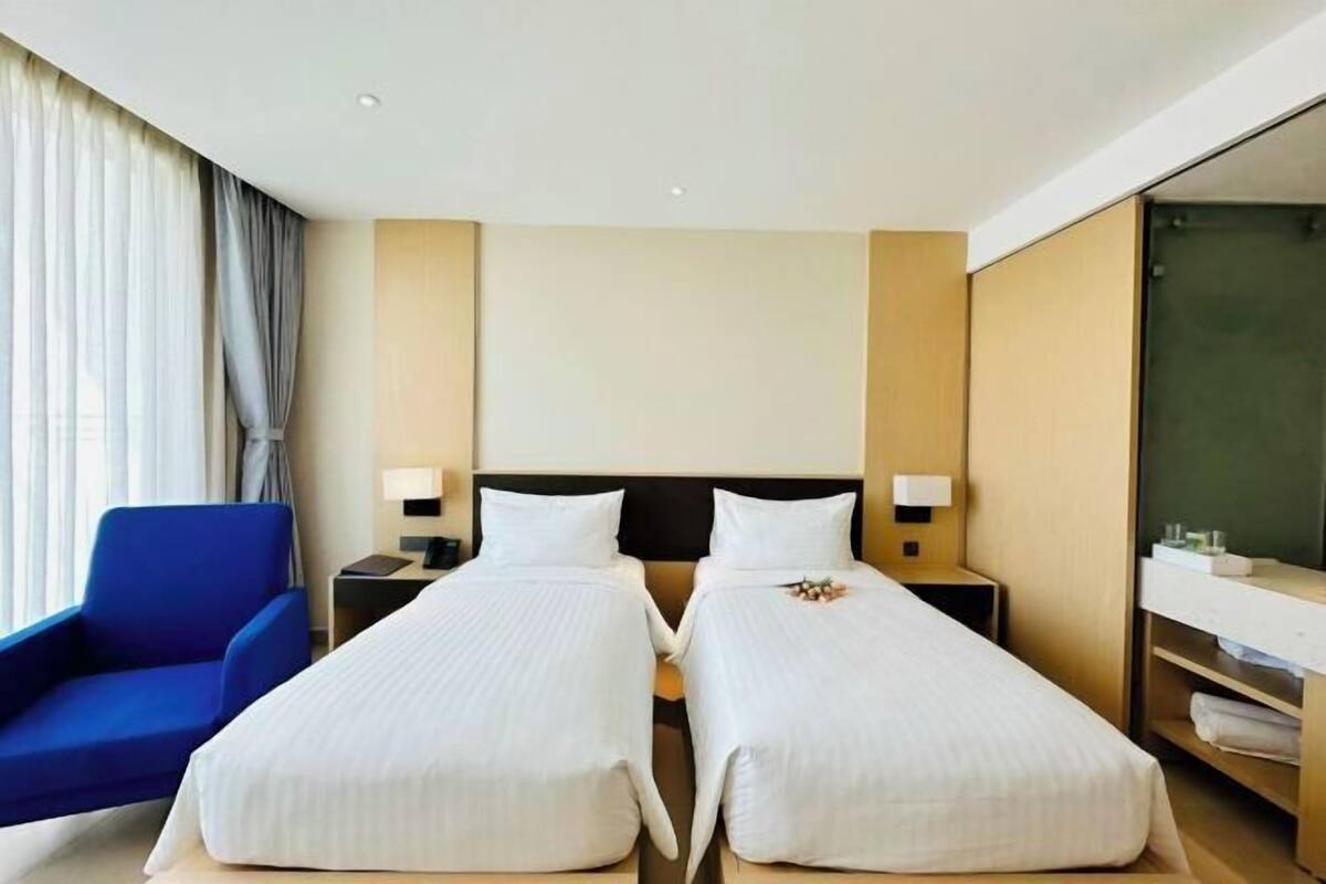Une chambre d’hôtel avec deux lits, une chaise bleue, une table de chevet avec une lampe, un miroir et une grande fenêtre avec des rideaux.