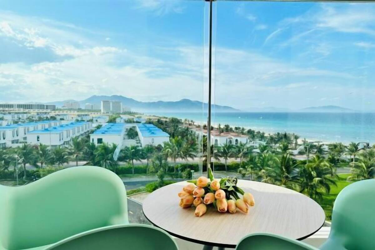 Une chambre d’hôtel avec vue sur la plage et les montagnes, une table ronde ornée d’un bouquet de fleurs et des chaises vertes.