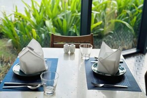 A table set with a folded napkin, a glass, and cutlery, with a view of green foliage through a window. - The Cam Ranh Oceanview (Cam Ranh)