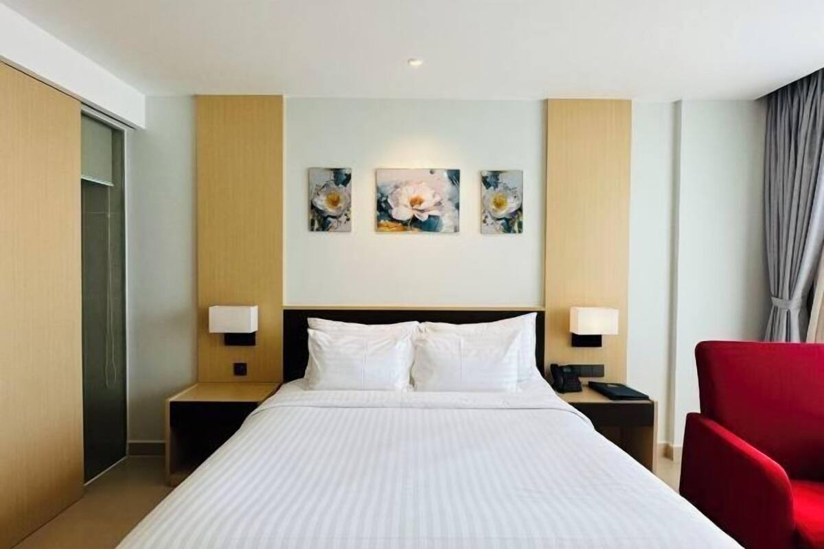 Une chambre d’hôtel avec un grand lit, deux tables de chevet, un fauteuil rouge et trois tableaux représentant des motifs floraux accrochés au mur.