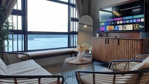A room with a large window overlooking the sea, a flat-screen TV mounted on the wall, and a round wooden table with a vase of flowers. - JESSELTON QUAYFULL SEA VIEWHIGH FLOORCOMFORT2BR4 PAX (Kota Kinabalu)