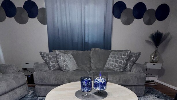 Een woonkamer met een ronde salontafel, een grijze bank, gekleurde kussens en een blauw-wit tapijt met dessin.
