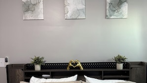 Een slaapkamer met een bed, drie abstracte kunstwerken aan de muur, een donker hoofdeinde, twee kleine potplanten en twee decoratieve kussens met tekst.