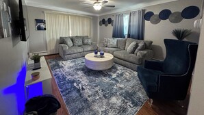 Een moderne woonkamer met een grijze zithoek, een ronde salontafel, een blauwe fauteuil en een plafondventilator.